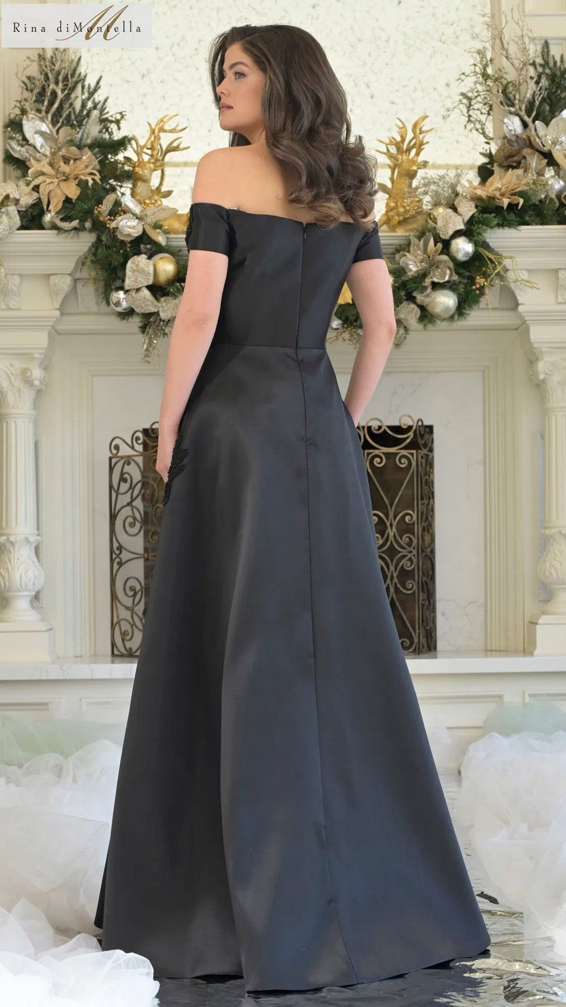 A woman with wavy brown hair wears the Rina Di Montella RD3344 long formal dress, standing indoors before a fireplace decorated with greenery and gold accents.